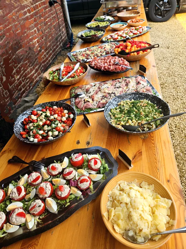 Een lange houten tafel vol schotels: gevulde tomaatjes met ei, tomaat met mozzarella, pastasalade met dille, carpaccio, rauwe ham, salades en een kom Parmezaanschilfers.