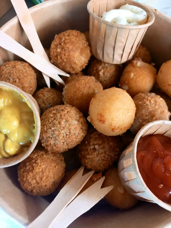 Een kartonnen kom vol aperitiefkroketjes en kaasballetjes, met mosterd, ketchup en tartaar in papieren potjes en houten vorkjes.