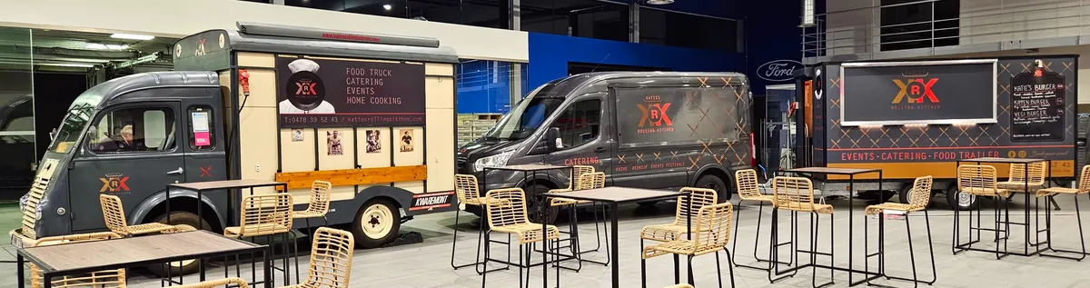 De Goelette, de Transit en de trailer 's avonds samen in een showroom, met statafels en rieten stoelen ervoor.